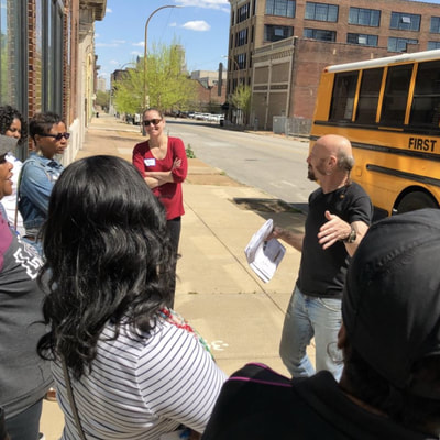 man speaking to group on sidewalk by school bus