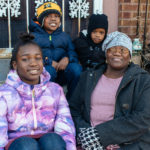 black woman and three smiling black children all in coats sitting on front steps