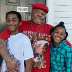 black man with his arms around two black children on front porch