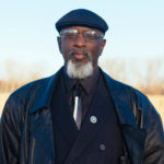 confidently smiling goateed black man with cap and glasses